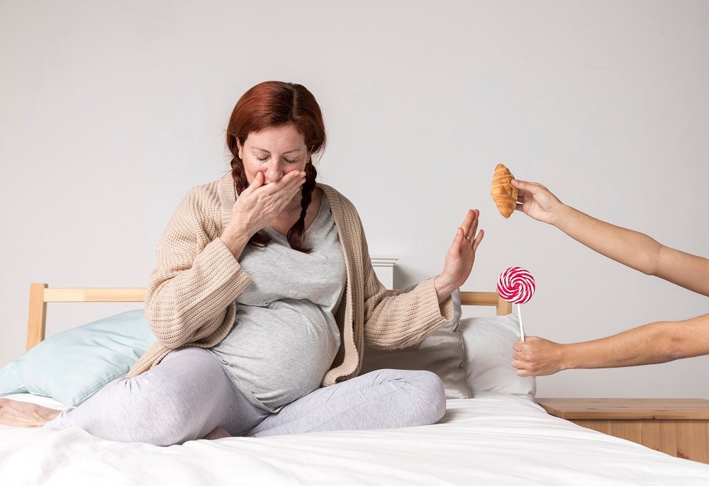 Pregnant woman experiencing morning sickness while refusing unhealthy food options like a croissant and candy.